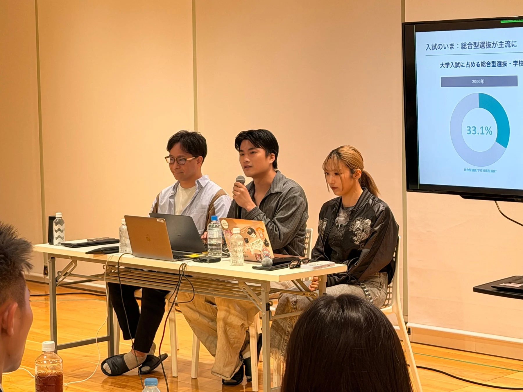 3人の登壇者がテーブルに着席し、聴衆を前にプレゼンテーションを行っている様子