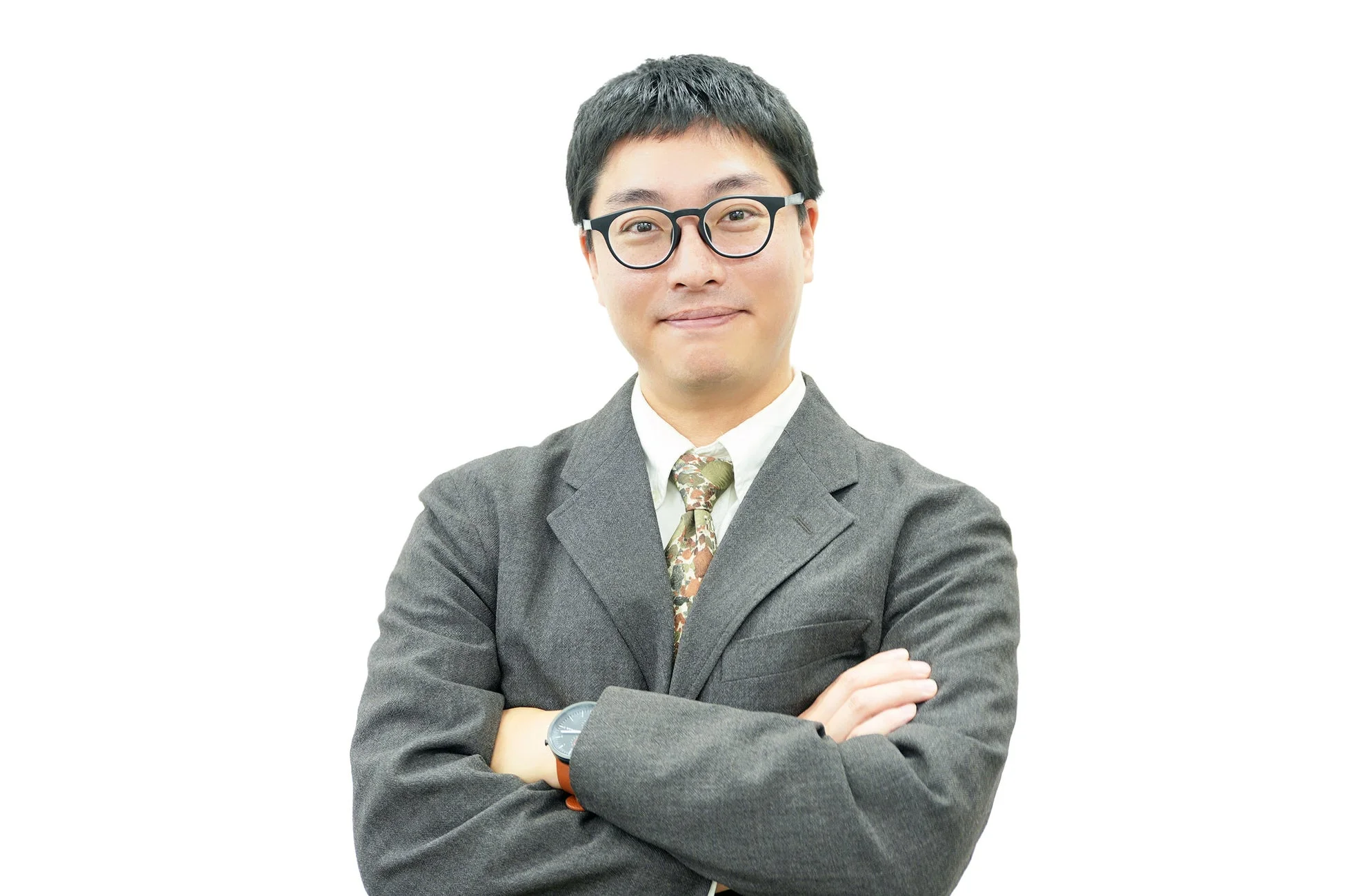 白い背景の前で、眼鏡をかけたアジア系の男性がスーツを着て腕を組み、笑顔でカメラを見ているポートレート写真です。