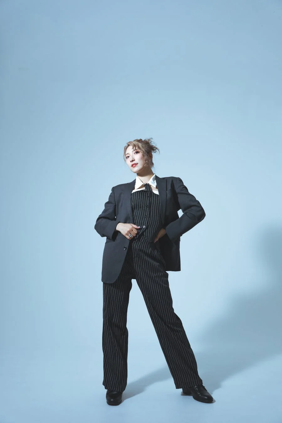 明るい青の背景を背に、ストライプのスーツをスタイリッシュに着こなした若い女性の全身ポートレート