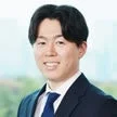 ビジネススーツを着た若いアジア人男性が、明るくぼやけた背景の前でカメラに向かって笑顔を見せているポートレート写真です。