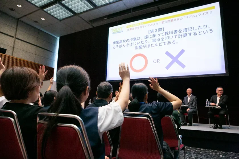 「都立商業高校のリアル」クイズイベントの様子。聴衆がスクリーンに映し出された授業に関する問題に対し、挙手で回答している。壇上には2名の登壇者が座っている。