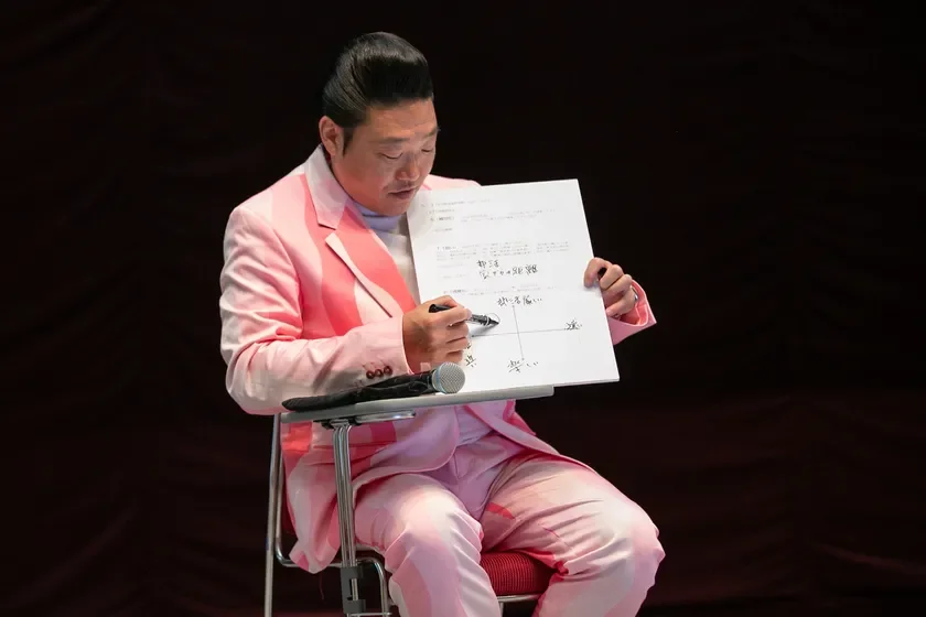 ピンクのスーツを着た男性が、ポンパドールヘアで椅子に座り、紙に書かれた師弟関係の距離や感情を示す図をペンで指しながら説明しています。講演やプレゼンテーションの場面のようです。