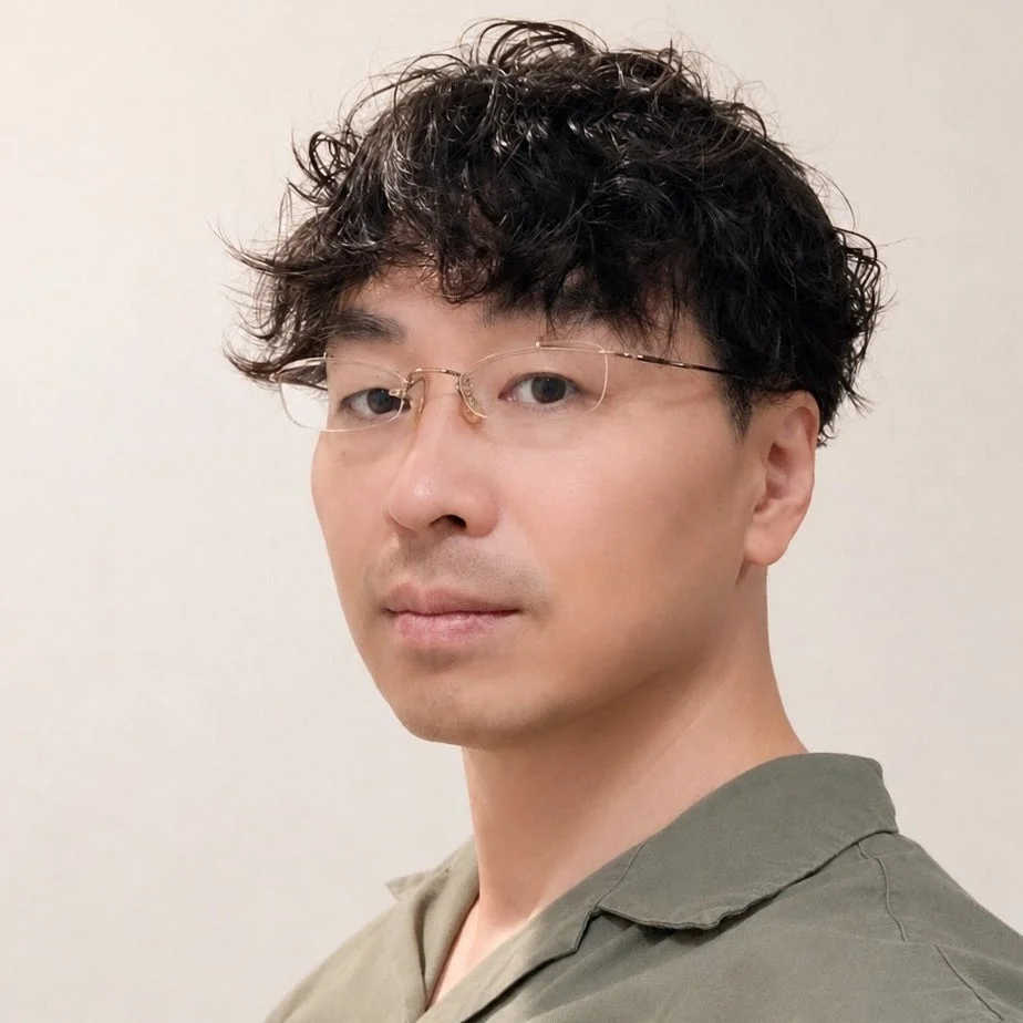 暗い色のカーリーヘアと眼鏡をかけた男性のポートレート写真です。緑色の襟付きシャツを着用し、まっすぐカメラを見つめています。背景は明るい無地です。