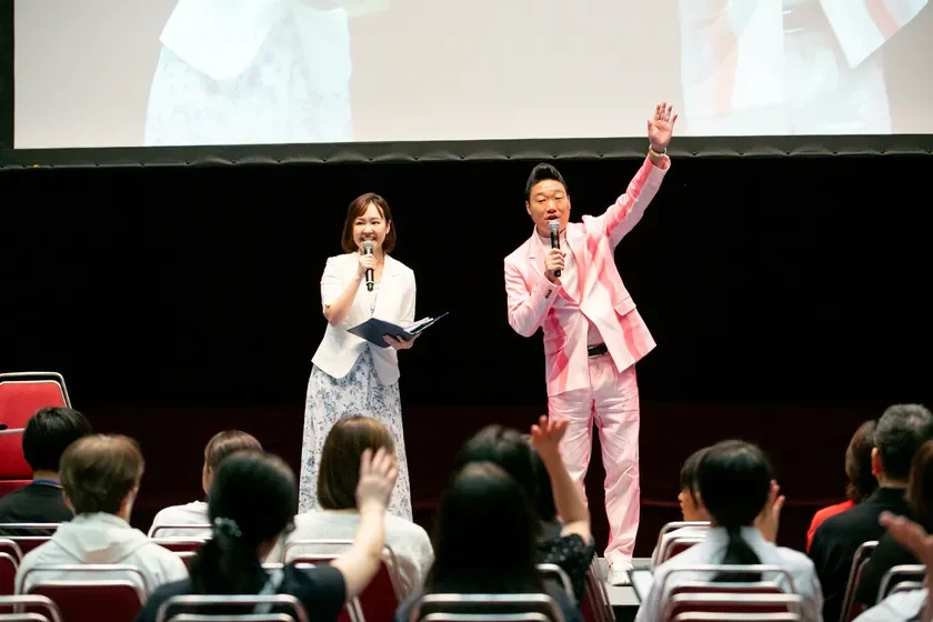 ステージ上で二人の登壇者が聴衆に向けて話しており、一部の聴衆は手を挙げています。男性はピンクのスーツ、女性は白のジャケットと花柄のスカートを着用しています。