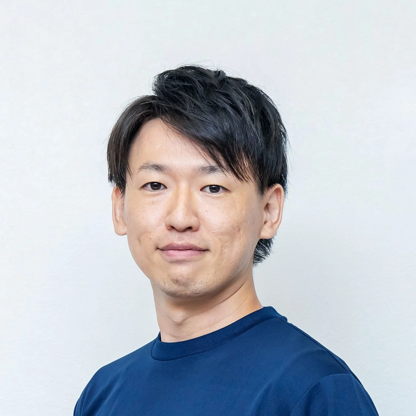 白い背景の前に立つ、紺色のTシャツを着たアジア人男性のバストアップポートレートです。カメラをまっすぐ見つめています。