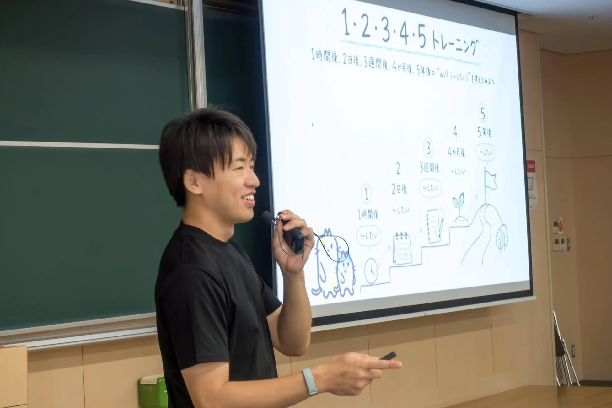 「1・2・3・4・5 トレーニング」と題された目標設定に関するスライドを背景に、笑顔の男性がマイクを手にプレゼンテーションを行っている。