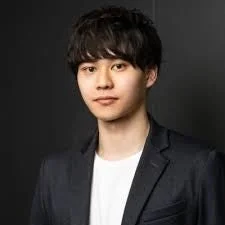 黒い背景の前で、白いTシャツの上にダークなジャケットを羽織った若い男性のポートレート