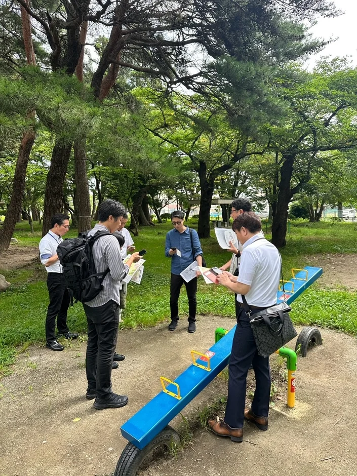 公園の木々の下で資料やスマートフォンを見ながら話し合っている複数の男性