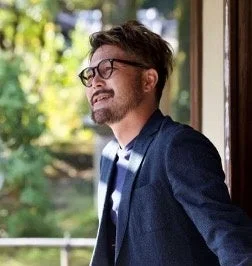 眼鏡と髭を生やした男性が、屋外の緑豊かな背景を背に、横を向いて穏やかな表情