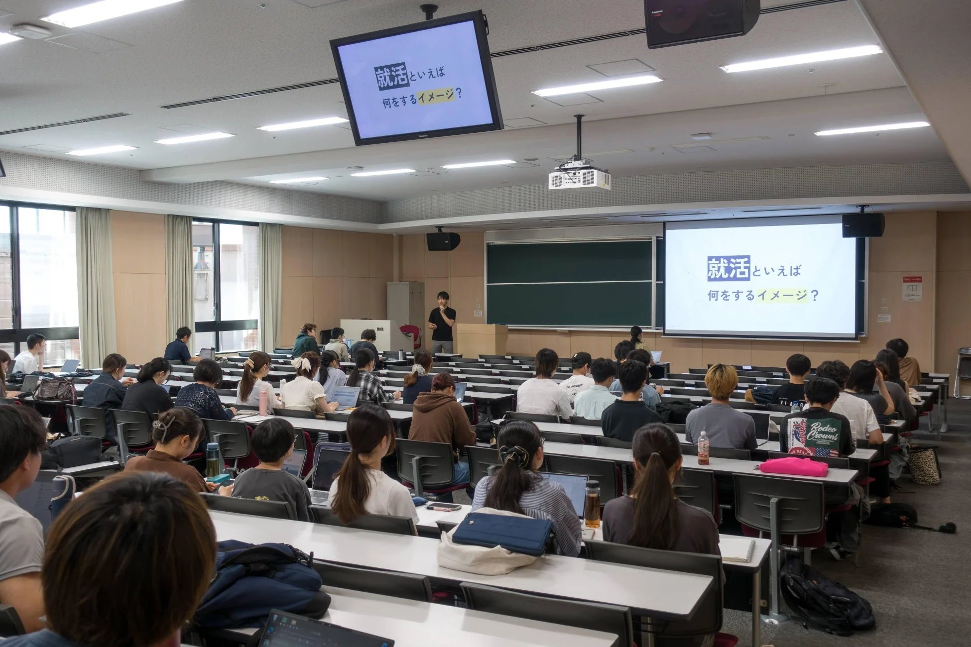 大学の講義室で、講師が学生たちにプレゼンテーションを行っている。スクリーンには「就活 といえば 何をするイメージ?」というテキストが表示されており、就職活動に関する講義やセミナーの様子がうかがえる。多くの学生がノートパソコンを使用している。
