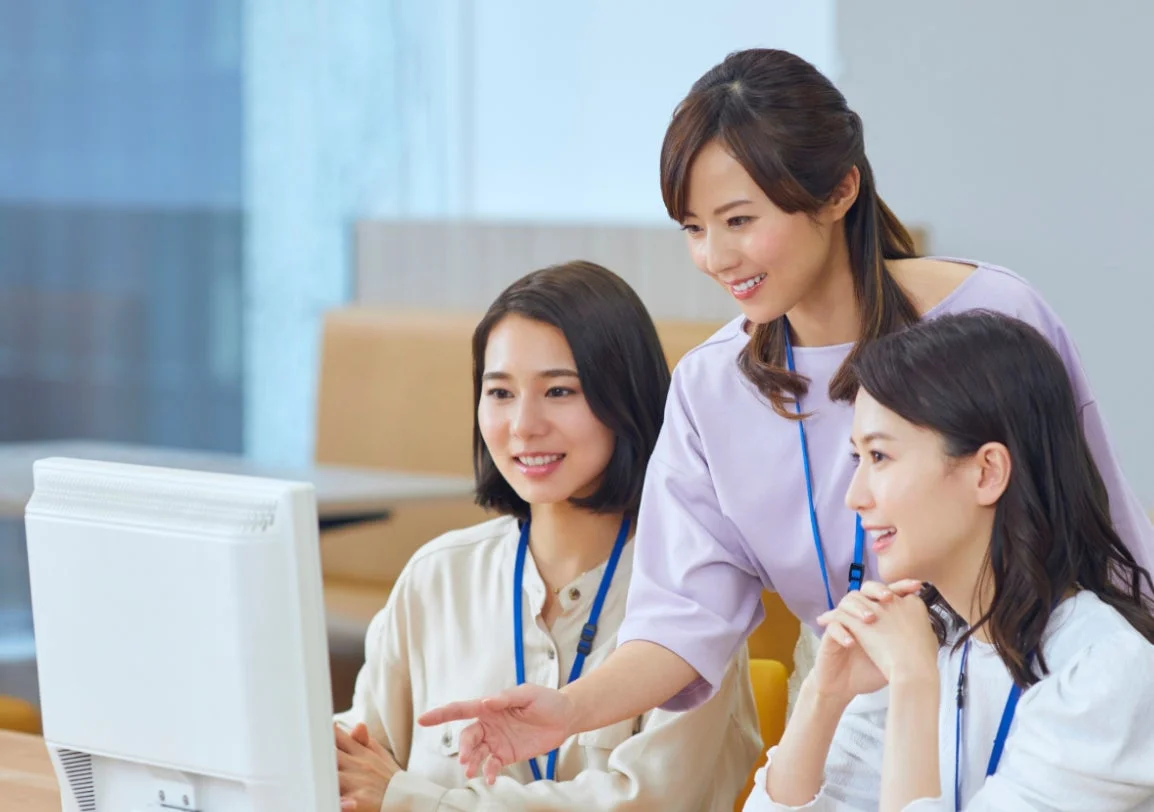 オフィスで3人の女性がPC画面を見ながら笑顔で協力し合っている様子